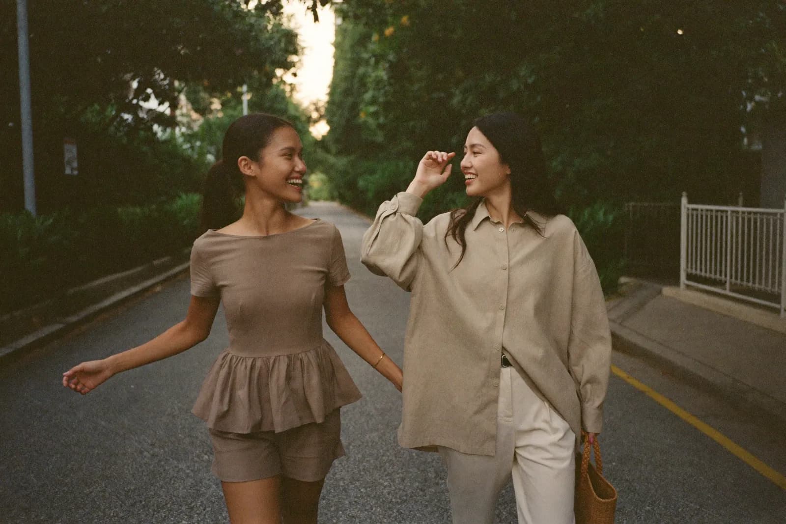 Two friends laughing on a tree-lined street, one in a ruffle-hem set, one in an oversized linen shirt